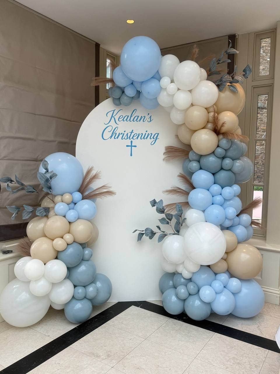 Balloon arch decoration with blue, white, and beige balloons for a christening event.