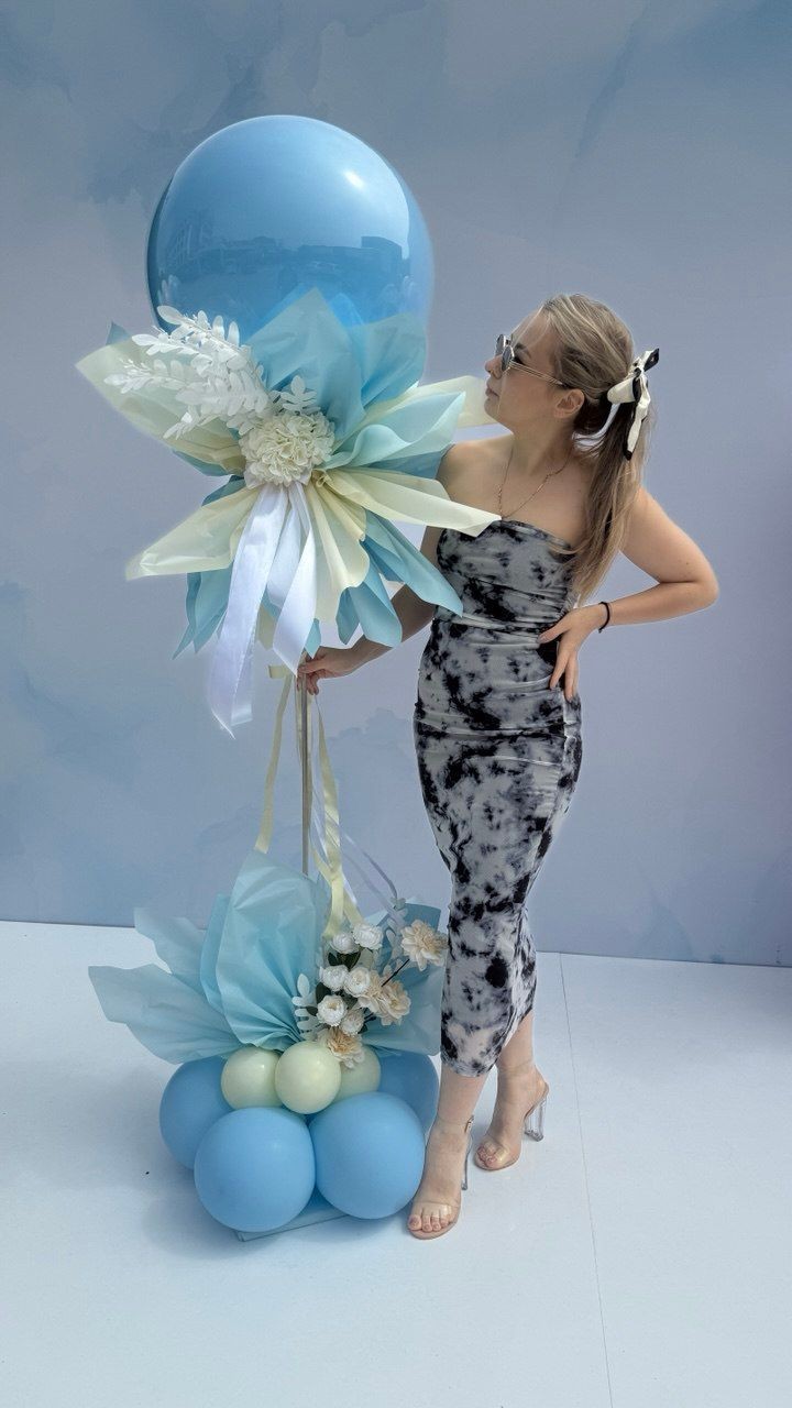 Woman in a patterned dress standing next to a large blue balloon display with flowers and ribbons.