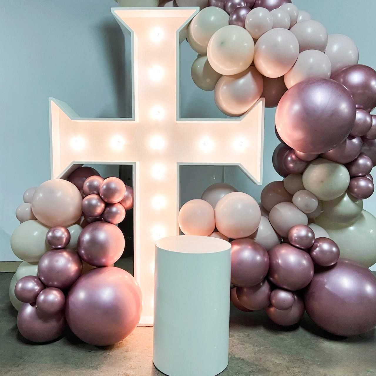 Illuminated cross sign surrounded by clusters of pink and beige balloons.